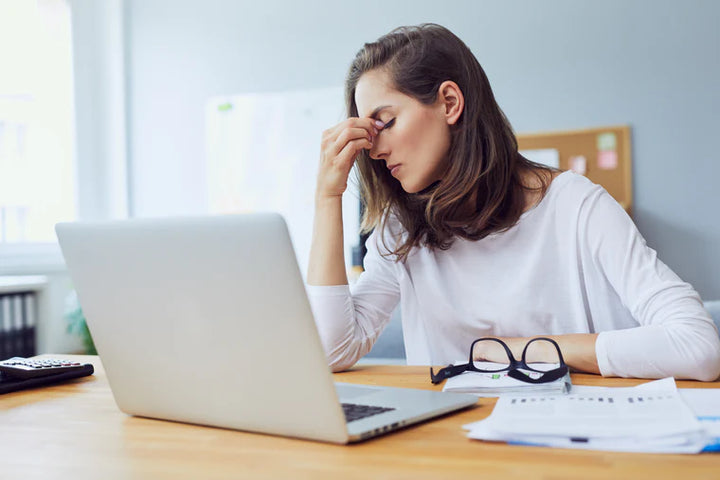 woman sitting at a desk in the day time with a laptop and paperwork in front of her. her glasses are on the table as well as she grabs the top of her nose indicating she has a headache