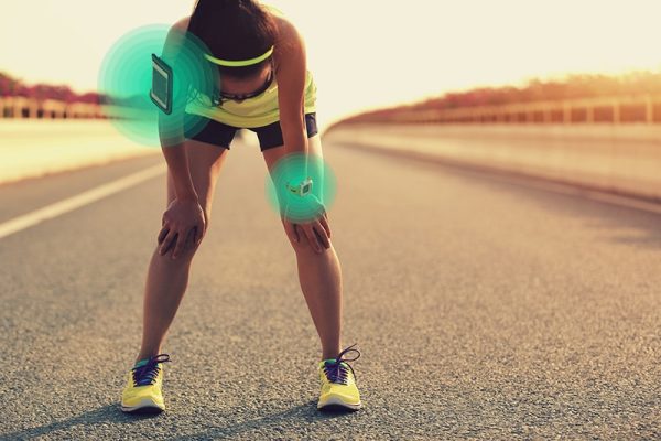 woman kneeling over with her hands on her knees taking a break from running. She is wearing athletic wear, she is on a paved road and has a cell phone and a fitness tracker.