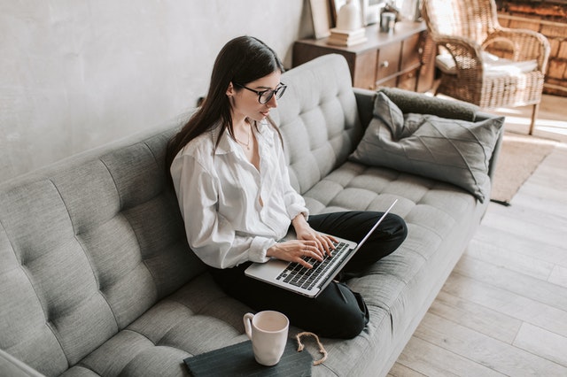woman sitting on couch, criss cross with a laptop in her lap. She appears to be drinking coffee and working in a living room.